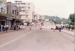 Uma fotografia de um desfile ou evento público em uma rua urbana, possivelmente em uma cidade brasileira como Videira, onde um grupo carrega uma faixa referente à 'XXII Convenção Distrito L-23'. A cena mostra pessoas caminhando pela via, edifícios comerciais ao fundo e elementos típicos de um evento cívico ou associativo, provavelmente ocorrido entre as décadas de 1980 e 1990.