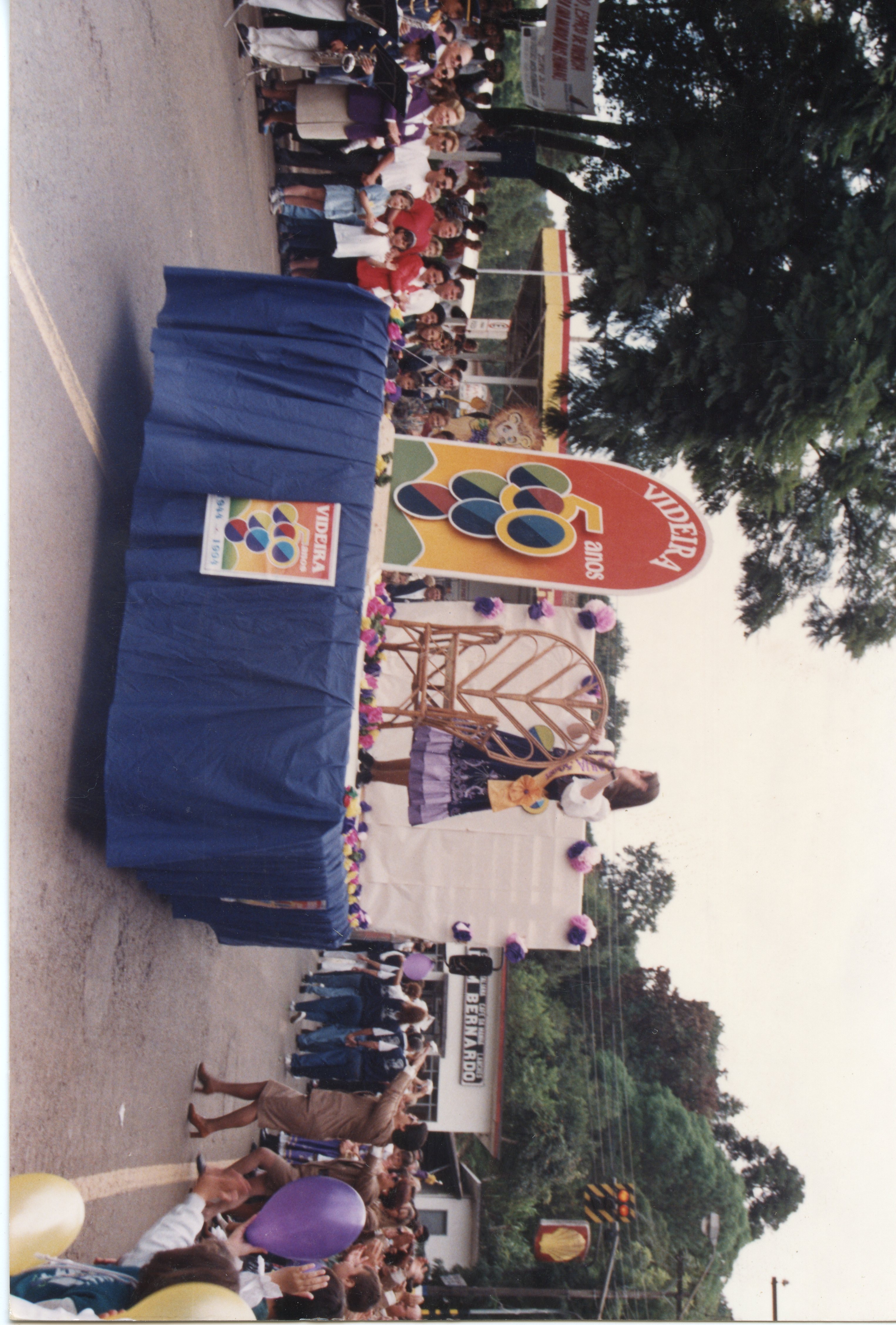 Fotografia de um desfile comemorativo em Videira, celebrando os 50 anos da cidade. O carro alegórico exibe o logotipo do cinquentenário e uma pessoa vestida com trajes típicos, em um contexto de celebração pública ao ar livre.
