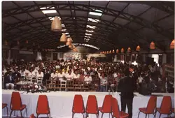 Uma fotografia de um grande evento social ou convenção, possivelmente um encontro de Lions Clube, realizado em um amplo galpão com estrutura metálica. A imagem mostra uma multidão sentada em cadeiras dispostas em fileiras, com uma mesa de honra em destaque e iluminação pendente característica, sugerindo um evento ocorrido em meados da década de 1990.