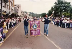 Uma fotografia de um desfile cívico ou evento comunitário em Videira, Santa Catarina, onde membros do Lions Clube carregam um estandarte. A cena mostra uma rua urbana com pessoas alinhadas nas calçadas, balões coloridos e vestimentas típicas do final do século XX.