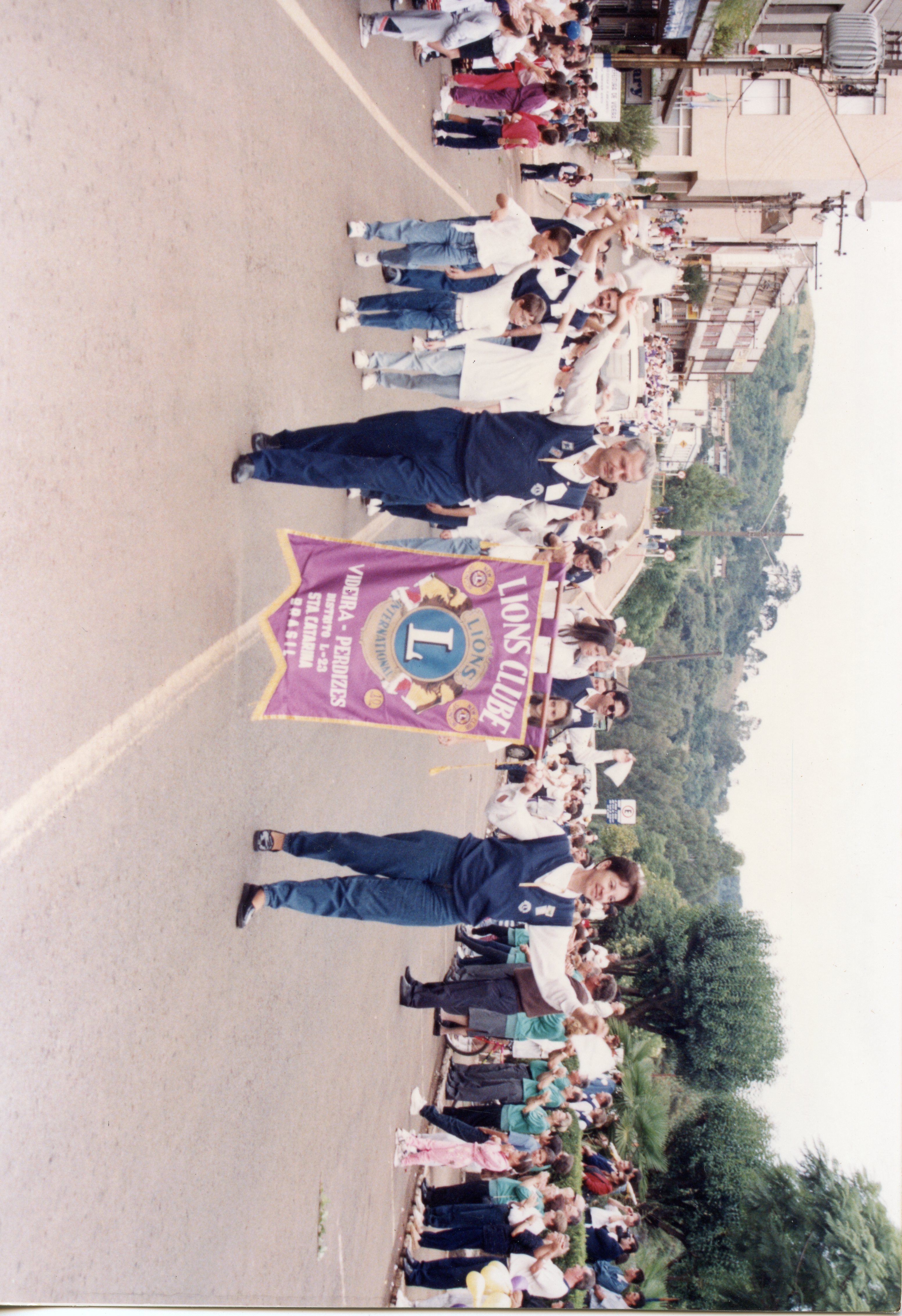 Uma fotografia de um desfile cívico ou evento comunitário em Videira, Santa Catarina, destacando membros do Lions Clube carregando uma bandeira oficial. A cena mostra um grupo de pessoas caminhando em uma rua urbana, com vestimentas casuais típicas do final do século XX, sugerindo um evento social ou comemorativo local.