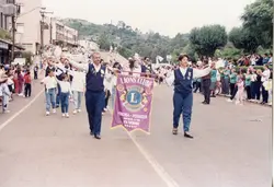 Uma fotografia de um desfile cívico ou evento comunitário em Videira, Santa Catarina, destacando membros do Lions Clube carregando uma bandeira oficial. A cena mostra um grupo de pessoas caminhando em uma rua urbana, com vestimentas casuais típicas do final do século XX, sugerindo um evento social ou comemorativo local.
