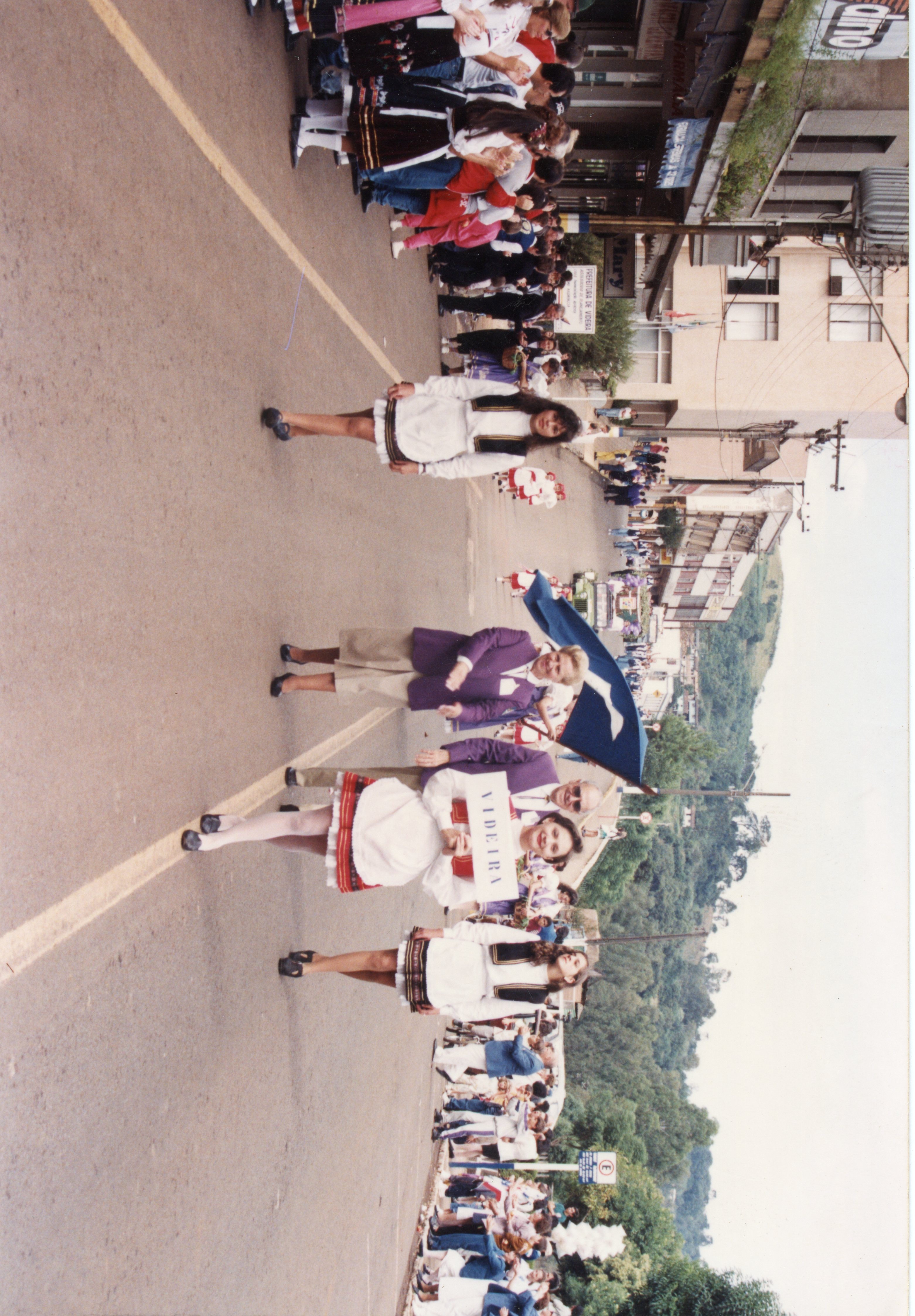 Uma fotografia de um desfile cívico ou cultural na cidade de Videira, Santa Catarina, provavelmente nas décadas de 1980 ou 1990. Pessoas vestidas com trajes típicos europeus caminham pela rua central, carregando uma placa com o nome da cidade, enquanto espectadores observam das calçadas.