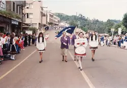 Uma fotografia de um desfile cívico ou cultural na cidade de Videira, Santa Catarina, provavelmente nas décadas de 1980 ou 1990. Pessoas vestidas com trajes típicos europeus caminham pela rua central, carregando uma placa com o nome da cidade, enquanto espectadores observam das calçadas.
