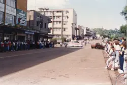 Uma cena de rua urbana em uma cidade brasileira, possivelmente durante um desfile ou evento cívico, com uma multidão acompanhando uma faixa do Lions Clube. A arquitetura dos prédios e as vestimentas sugerem o final do século XX, provavelmente entre as décadas de 1980 e 1990.