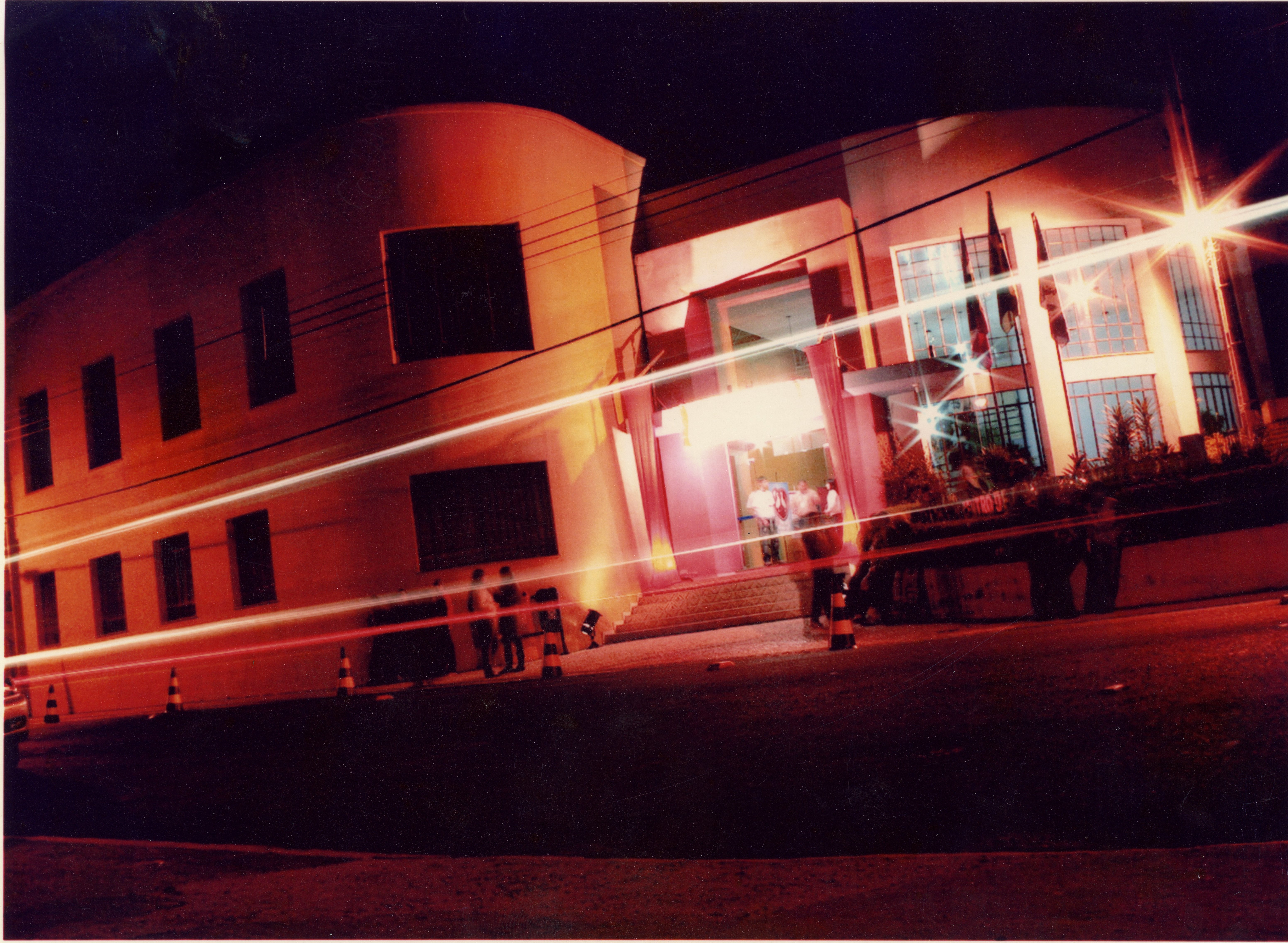 Fotografia noturna de um edifício público com arquitetura de linhas curvas, possivelmente uma prefeitura ou centro cultural, iluminado por luzes artificiais. O efeito de longa exposição cria rastros de luz horizontais, sugerindo o movimento de veículos em frente ao local, com algumas pessoas visíveis na entrada.