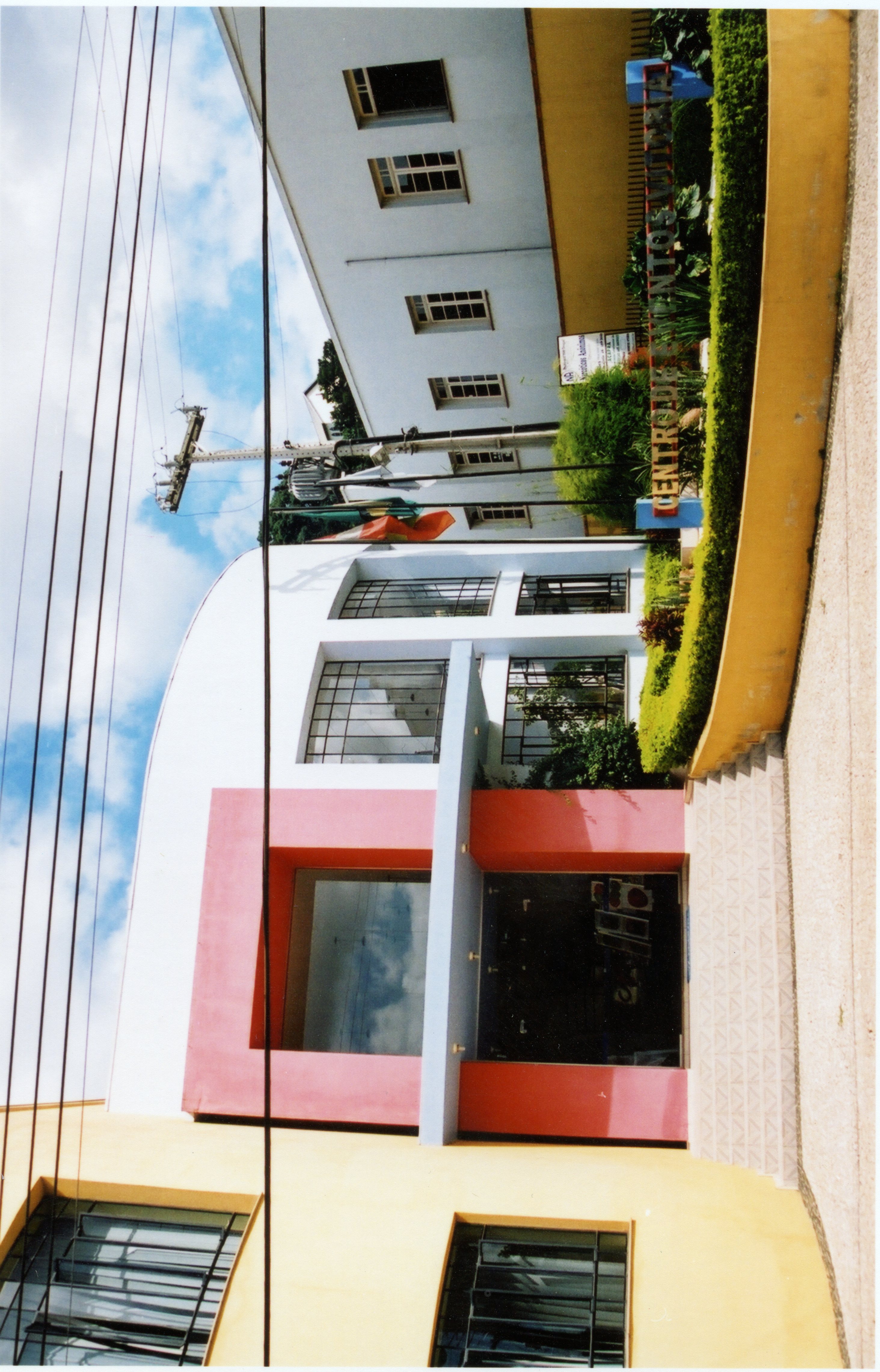 Fotografia colorida de um edifício comercial moderno com fachada pintada em tons de amarelo, branco e rosa, localizado em uma área urbana. O letreiro indica 'Centro de Eventos Videira', sugerindo um espaço público ou cultural na cidade de Videira, Santa Catarina.