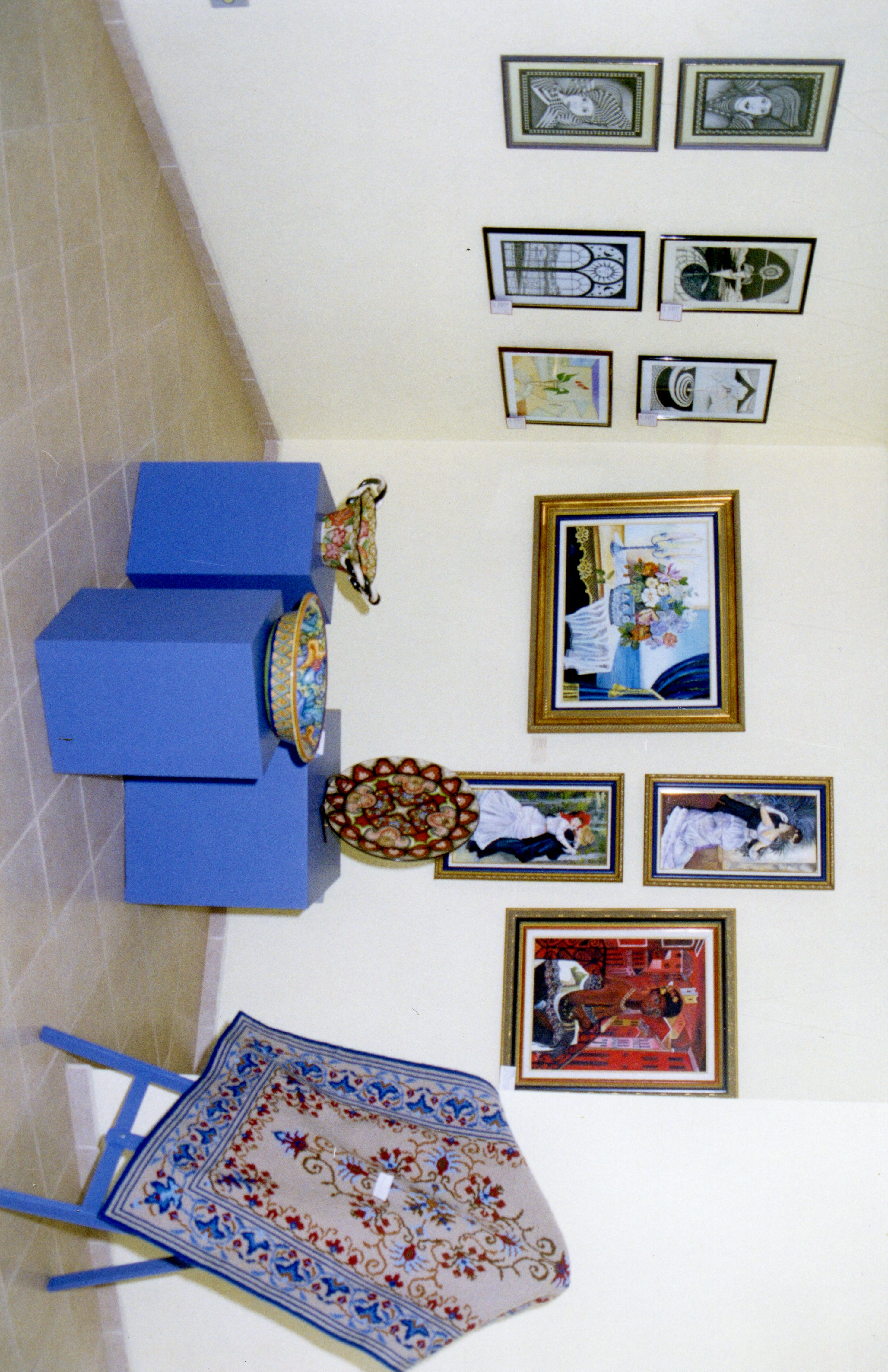 Uma exposição de arte em um ambiente interno, apresentando diversas pinturas emolduradas dispostas na parede e objetos decorativos, como pratos pintados, sobre pedestais azuis. O espaço parece ser uma galeria ou centro cultural, com um tapete decorativo em primeiro plano, sugerindo um evento de exibição artística contemporânea.