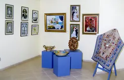Uma exposição de arte em um ambiente interno, apresentando diversas pinturas emolduradas dispostas na parede e objetos decorativos, como pratos pintados, sobre pedestais azuis. O espaço parece ser uma galeria ou centro cultural, com um tapete decorativo em primeiro plano, sugerindo um evento de exibição artística contemporânea.