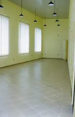Uma sala interna vazia com paredes pintadas de amarelo claro e um teto com textura quadriculada. O ambiente possui três janelas com persianas verticais brancas e várias luminárias de chão minimalistas espalhadas pelo piso, sugerindo um espaço comercial ou de exposição.