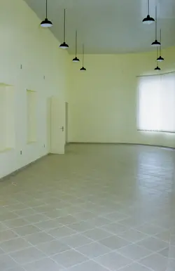 Uma sala ampla e vazia com paredes pintadas em tom claro, piso de cerâmica e iluminação pendente moderna. O ambiente parece ser um espaço comercial ou institucional contemporâneo, caracterizado pela simplicidade e pela ausência de mobília.