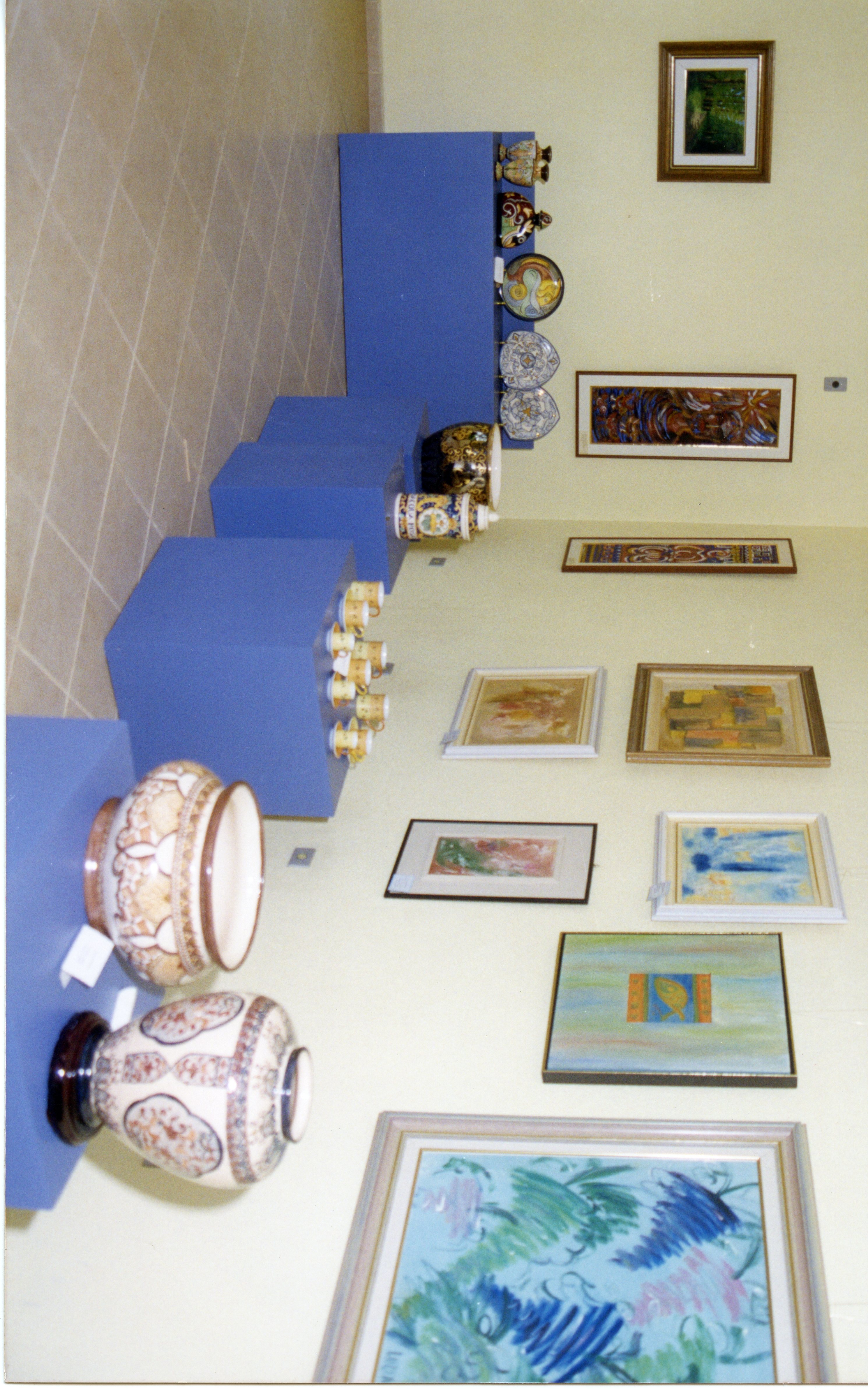 Uma exposição de arte e cerâmica em um ambiente interno, possivelmente uma galeria ou museu local. O espaço apresenta pedestais azuis exibindo vasos e xícaras decoradas, enquanto as paredes estão adornadas com diversas pinturas emolduradas, sugerindo um evento cultural ou mostra artística.