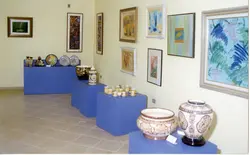 Uma exposição de arte e cerâmica em um ambiente interno, possivelmente uma galeria ou museu local. O espaço apresenta pedestais azuis exibindo vasos e xícaras decoradas, enquanto as paredes estão adornadas com diversas pinturas emolduradas, sugerindo um evento cultural ou mostra artística.