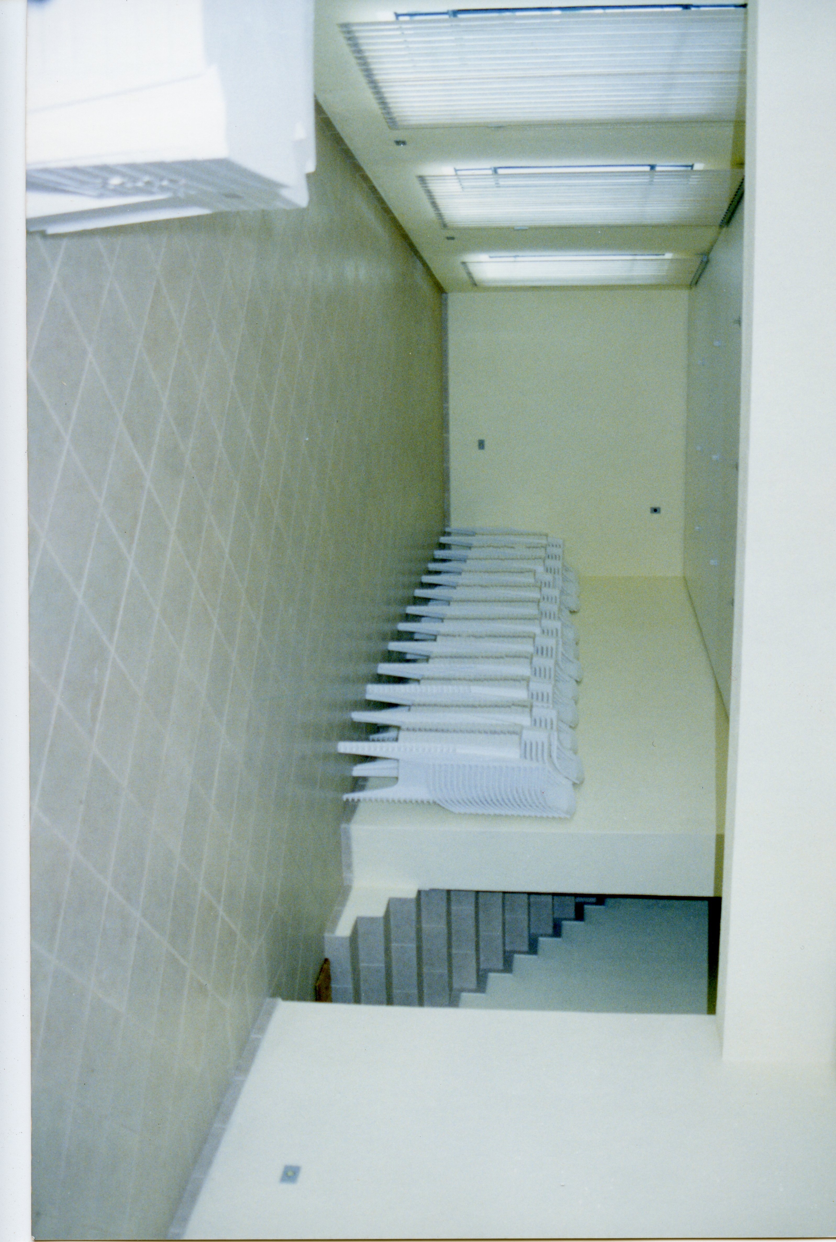 Uma imagem interna mostrando uma fileira de cadeiras de plástico empilhadas em um espaço arquitetônico moderno e minimalista. O ambiente apresenta paredes com revestimento em losangos e iluminação embutida no teto, sugerindo um salão de eventos ou área de circulação em um prédio público ou comercial contemporâneo.