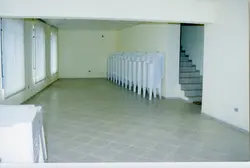 Uma imagem interna mostrando uma fileira de cadeiras de plástico empilhadas em um espaço arquitetônico moderno e minimalista. O ambiente apresenta paredes com revestimento em losangos e iluminação embutida no teto, sugerindo um salão de eventos ou área de circulação em um prédio público ou comercial contemporâneo.