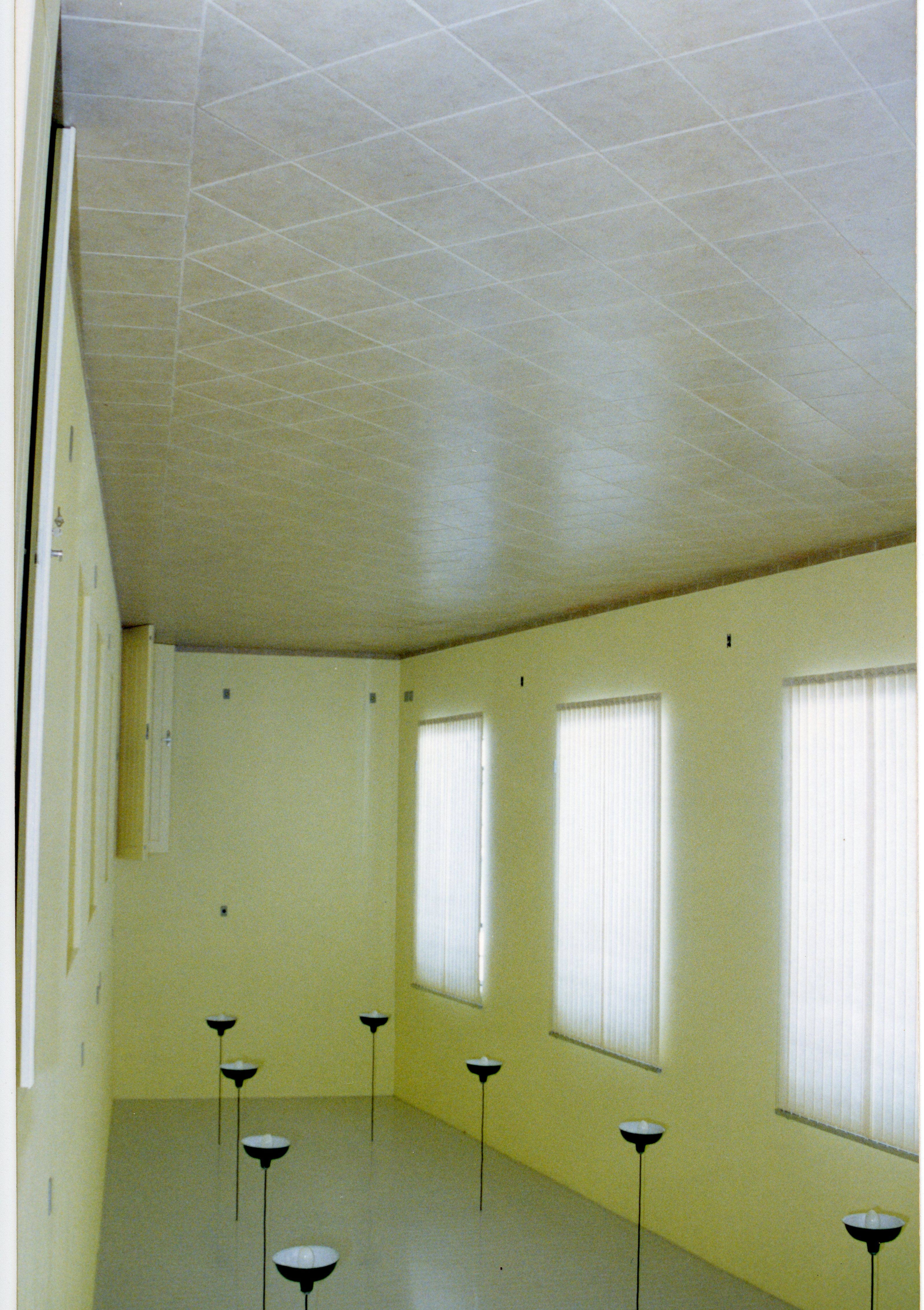 Uma sala interna vazia com paredes pintadas de amarelo claro e um teto com placas texturizadas. O ambiente apresenta várias luminárias de chão com cúpulas pretas e persianas verticais nas janelas, sugerindo um espaço comercial ou de escritório de meados da década de 1990.