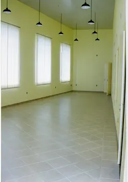 Uma sala interna vazia com paredes pintadas de amarelo claro e um teto com placas texturizadas. O ambiente apresenta várias luminárias de chão com cúpulas pretas e persianas verticais nas janelas, sugerindo um espaço comercial ou de escritório de meados da década de 1990.