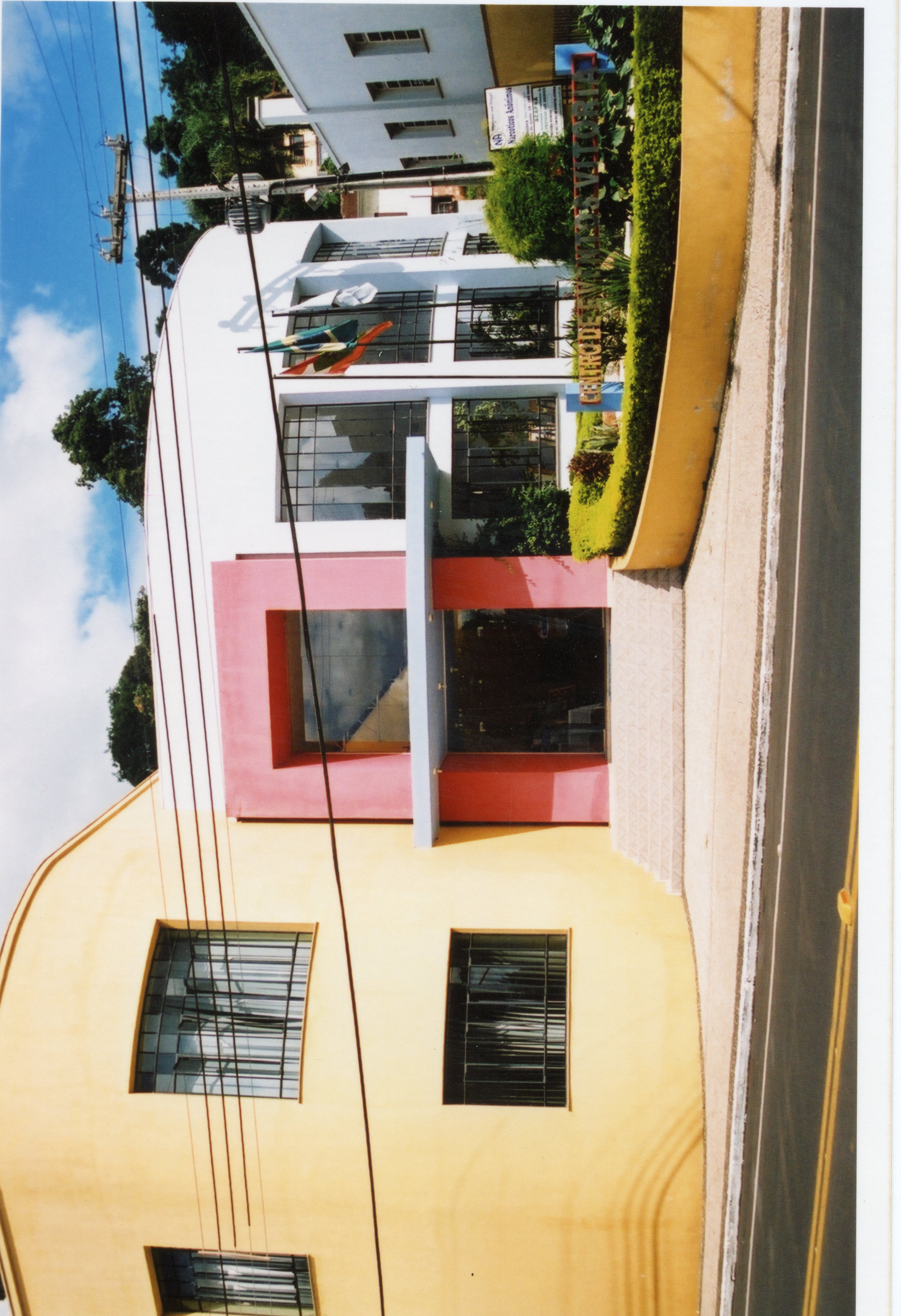 A imagem apresenta a fachada do Centro de Eventos Vitória, um edifício com arquitetura moderna e cores vibrantes como amarelo e rosa. O local parece ser um prédio público ou comercial em uma área urbana, possivelmente em uma cidade brasileira, com bandeiras hasteadas na entrada.