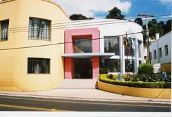 A imagem apresenta a fachada do Centro de Eventos Vitória, um edifício com arquitetura moderna e cores vibrantes como amarelo e rosa. O local parece ser um prédio público ou comercial em uma área urbana, possivelmente em uma cidade brasileira, com bandeiras hasteadas na entrada.