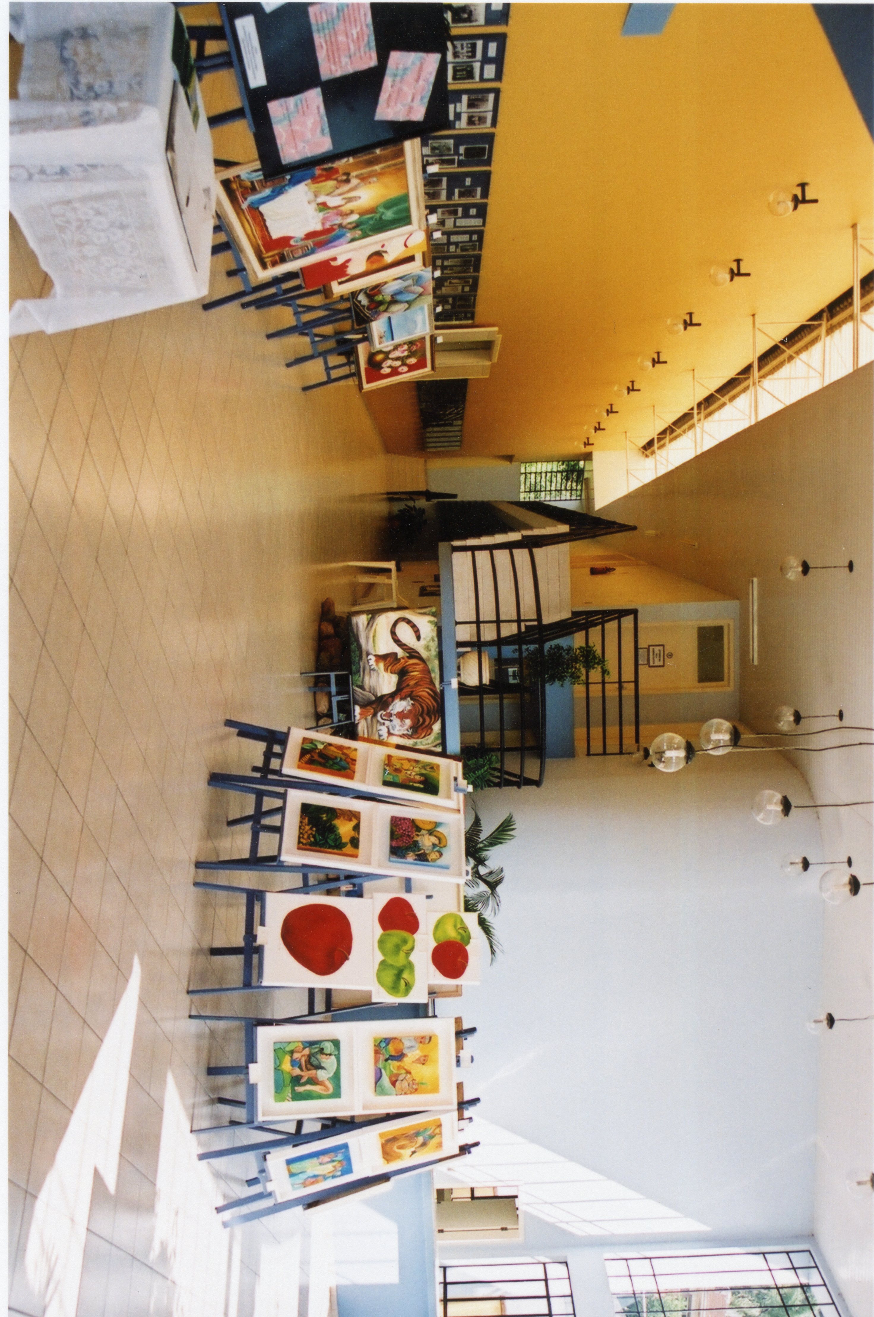 Uma exposição de arte organizada em um espaço interno moderno, possivelmente um centro cultural ou saguão de prédio público. Diversas pinturas estão dispostas em cavaletes, exibindo temas variados como naturezas-mortas e animais, em um ambiente com iluminação clara e arquitetura contemporânea.