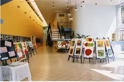 Uma exposição de arte organizada em um espaço interno moderno, possivelmente um centro cultural ou saguão de prédio público. Diversas pinturas estão dispostas em cavaletes, exibindo temas variados como naturezas-mortas e animais, em um ambiente com iluminação clara e arquitetura contemporânea.