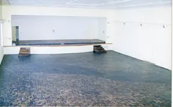 Uma fotografia de um espaço interior vazio, possivelmente uma galeria ou área comercial, com um piso de madeira em padrão espinha de peixe. O ambiente apresenta paredes brancas minimalistas e uma estrutura de escada de madeira, sugerindo uma arquitetura moderna ou contemporânea.