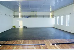 Uma visão interna de um espaço amplo e vazio, possivelmente um ginásio ou salão comunitário em fase de construção ou reforma. O ambiente apresenta um piso de madeira em uma lateral, paredes brancas e janelas com grades, sugerindo uma estrutura institucional ou pública típica de cidades do interior.