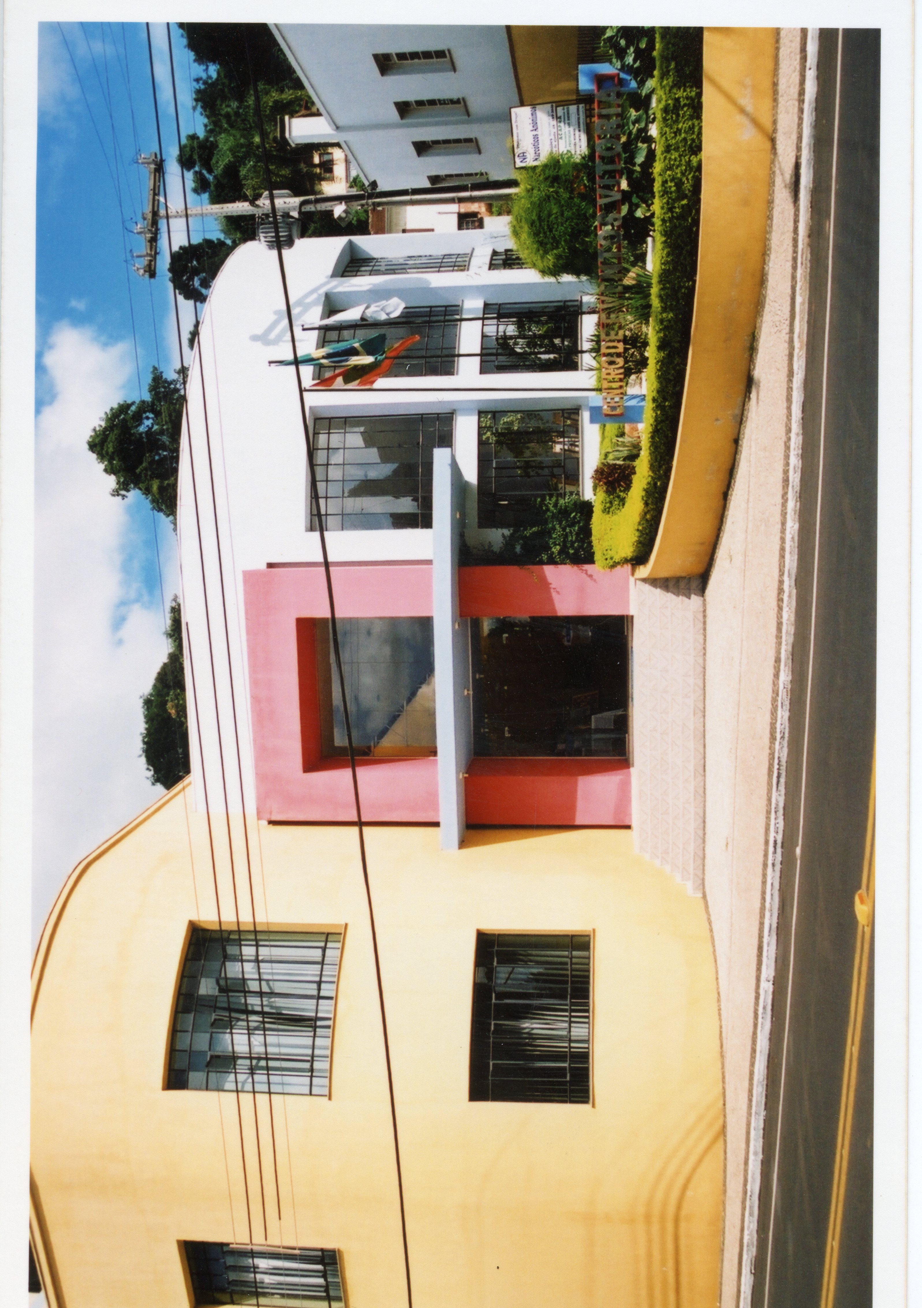 A imagem mostra a fachada do Centro de Eventos de Videira, um edifício com arquitetura moderna e cores vibrantes como amarelo e rosa. O local parece ser um espaço público ou institucional, capturado em um dia ensolarado com bandeiras hasteadas na entrada.