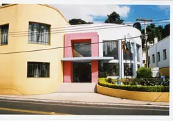 A imagem mostra a fachada do Centro de Eventos de Videira, um edifício com arquitetura moderna e cores vibrantes como amarelo e rosa. O local parece ser um espaço público ou institucional, capturado em um dia ensolarado com bandeiras hasteadas na entrada.