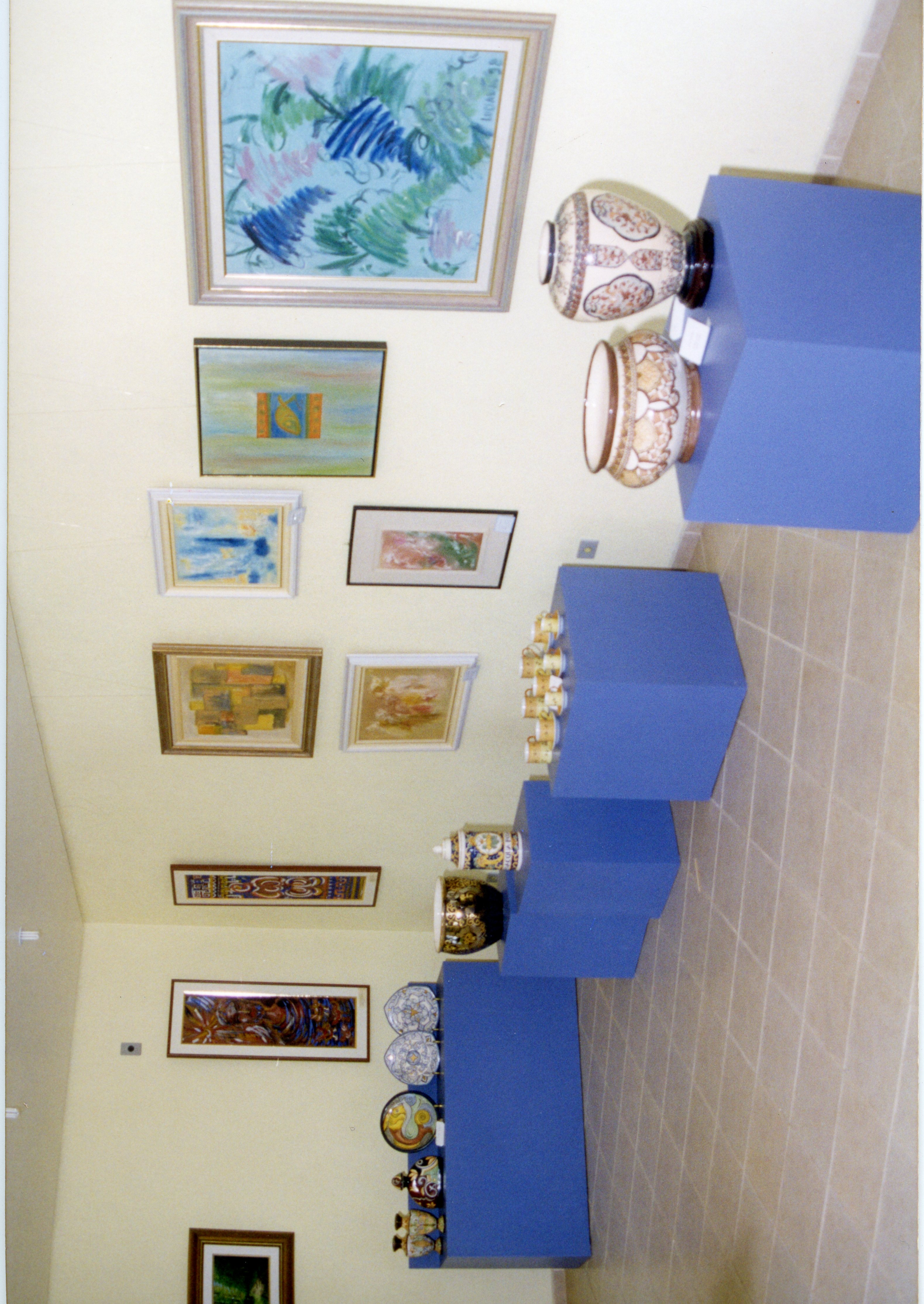 Uma exposição de arte e cerâmica em uma galeria ou espaço cultural. O ambiente apresenta quadros emoldurados nas paredes e peças de cerâmica decorativa dispostas sobre pedestais azuis.