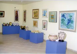 Uma exposição de arte e cerâmica em uma galeria ou espaço cultural. O ambiente apresenta quadros emoldurados nas paredes e peças de cerâmica decorativa dispostas sobre pedestais azuis.