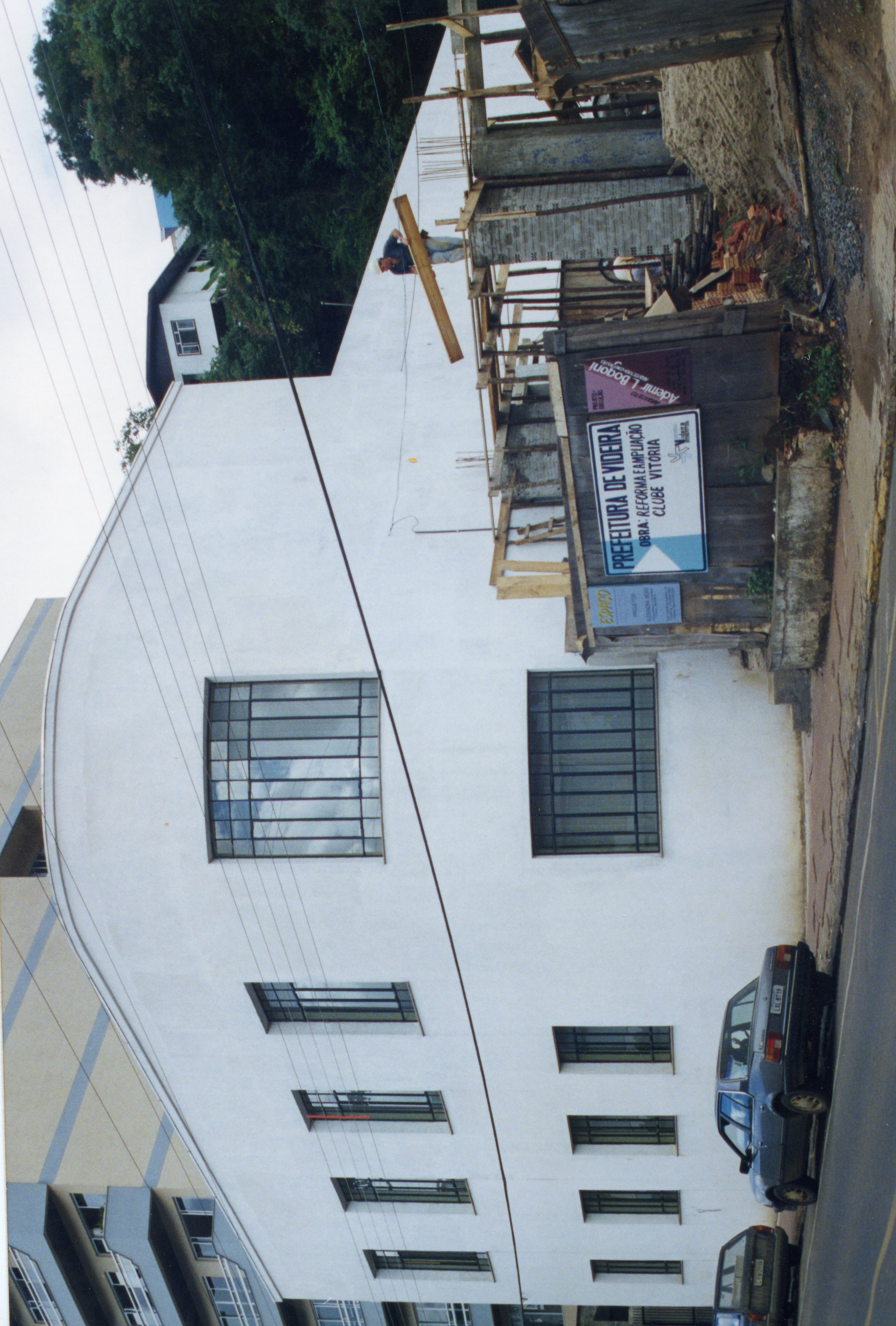 A imagem mostra uma obra de reforma e ampliação do Clube Vitória, em Videira, Santa Catarina, com uma placa da prefeitura visível no canteiro de obras. O edifício possui arquitetura funcionalista com janelas retangulares e está localizado em uma área urbana, com veículos estacionados na rua lateral.