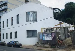 A imagem mostra uma obra de reforma e ampliação do Clube Vitória, em Videira, Santa Catarina, com uma placa da prefeitura visível no canteiro de obras. O edifício possui arquitetura funcionalista com janelas retangulares e está localizado em uma área urbana, com veículos estacionados na rua lateral.
