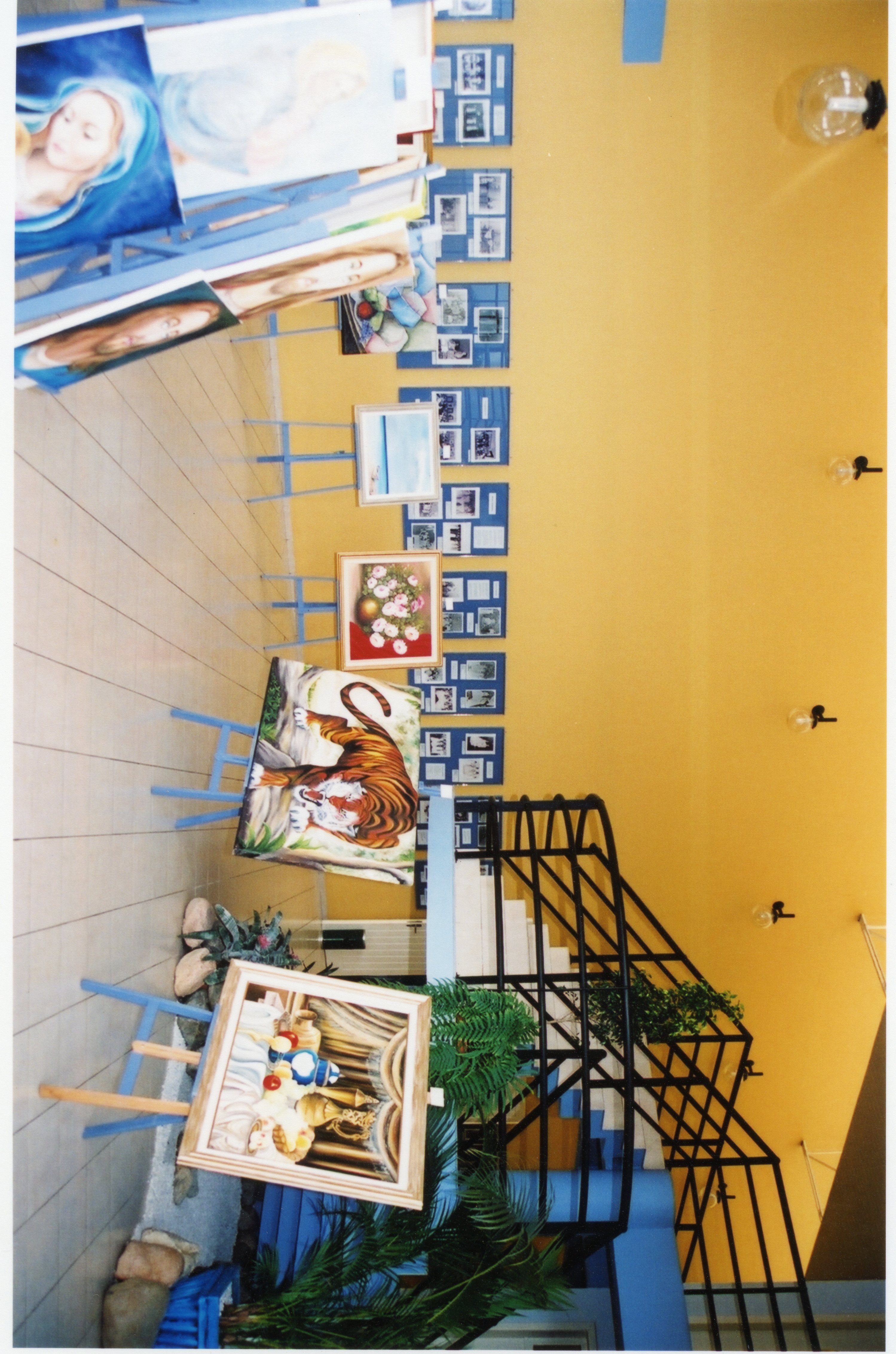 Uma exposição de arte organizada em um espaço interno com paredes amarelas, apresentando diversas pinturas em cavaletes dispostos em fila. Ao fundo, painéis azuis exibem fotografias, sugerindo uma mostra cultural ou histórica em um ambiente público ou galeria.