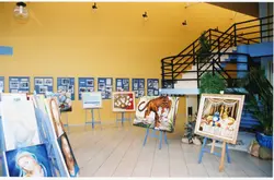 Uma exposição de arte organizada em um espaço interno com paredes amarelas, apresentando diversas pinturas em cavaletes dispostos em fila. Ao fundo, painéis azuis exibem fotografias, sugerindo uma mostra cultural ou histórica em um ambiente público ou galeria.
