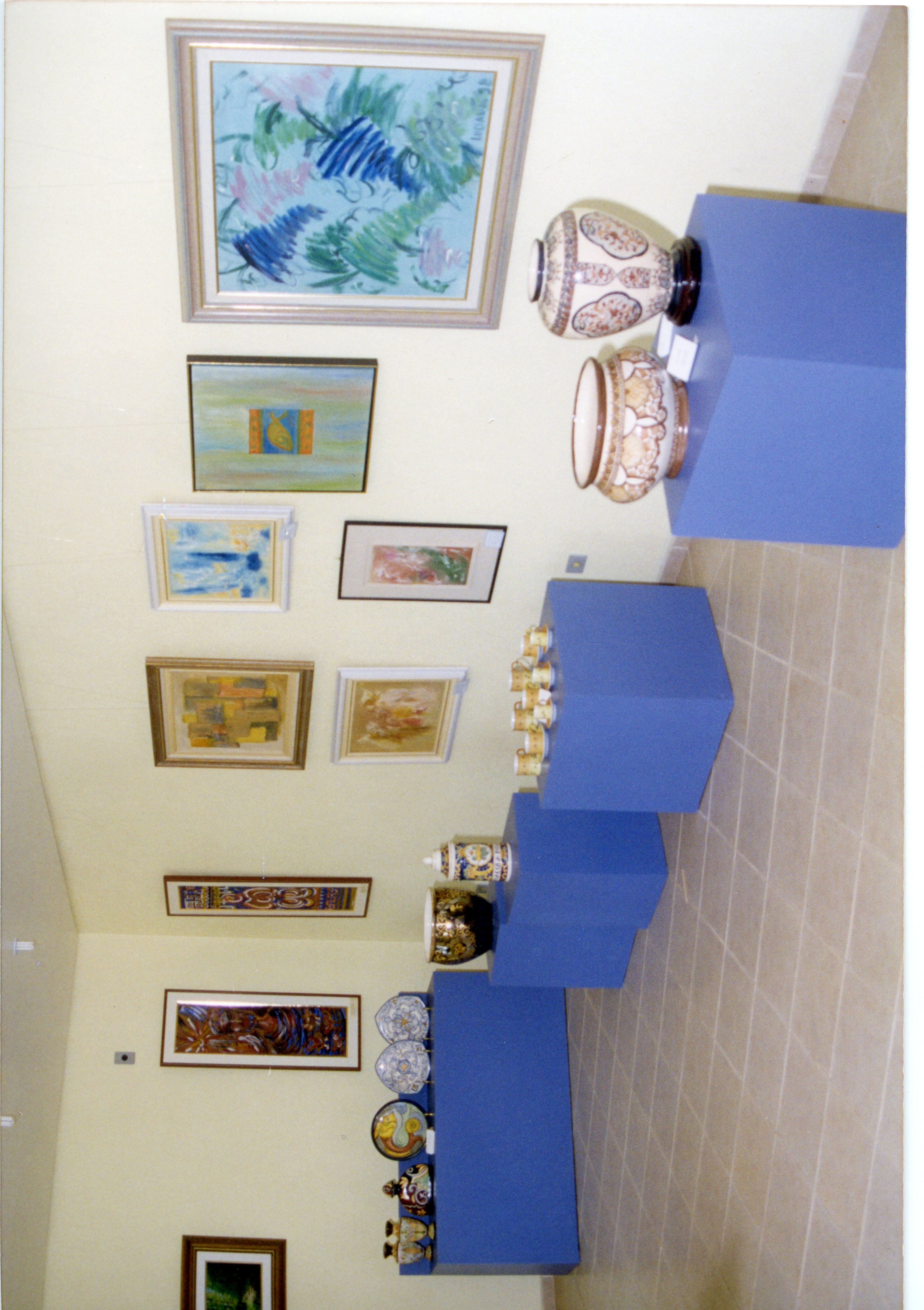 Uma exposição de arte organizada em uma galeria ou espaço cultural, exibindo diversas pinturas emolduradas nas paredes e cerâmicas decorativas dispostas sobre pedestais azuis. O ambiente apresenta uma estética típica de exposições de arte contemporânea ou de acervos municipais, possivelmente datando do final do século XX.
