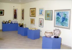 Uma exposição de arte organizada em uma galeria ou espaço cultural, exibindo diversas pinturas emolduradas nas paredes e cerâmicas decorativas dispostas sobre pedestais azuis. O ambiente apresenta uma estética típica de exposições de arte contemporânea ou de acervos municipais, possivelmente datando do final do século XX.