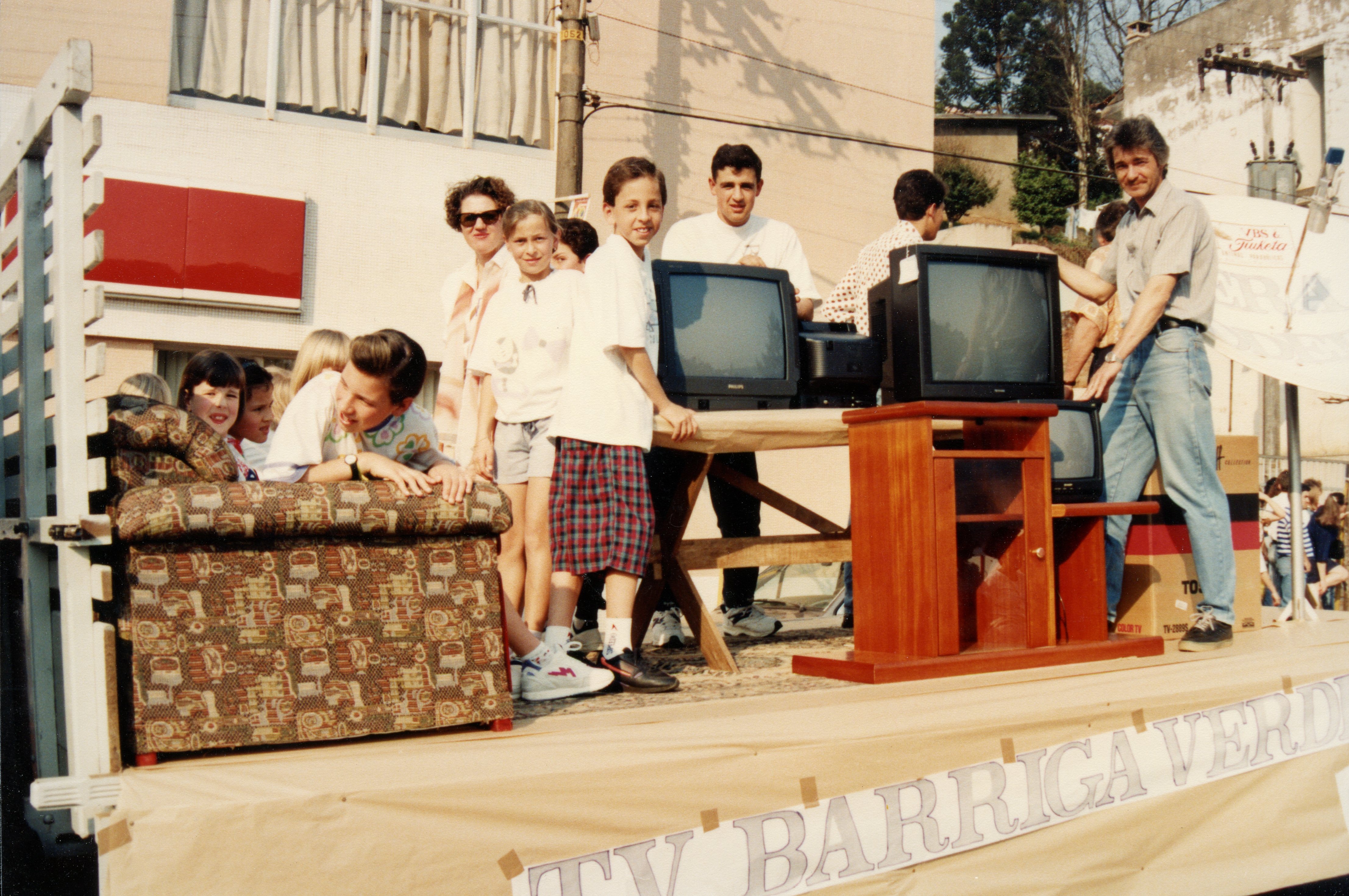 Uma fotografia colorida que mostra um grupo de pessoas, incluindo crianças e adultos, em cima de um caminhão decorado como um carro alegórico ou exibição promocional. O cenário inclui móveis, como um sofá estampado e um rack de madeira com televisores de tubo, sugerindo uma ação de marketing ou desfile comercial, possivelmente na década de 1990.