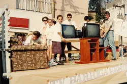 Uma fotografia colorida que mostra um grupo de pessoas, incluindo crianças e adultos, em cima de um caminhão decorado como um carro alegórico ou exibição promocional. O cenário inclui móveis, como um sofá estampado e um rack de madeira com televisores de tubo, sugerindo uma ação de marketing ou desfile comercial, possivelmente na década de 1990.
