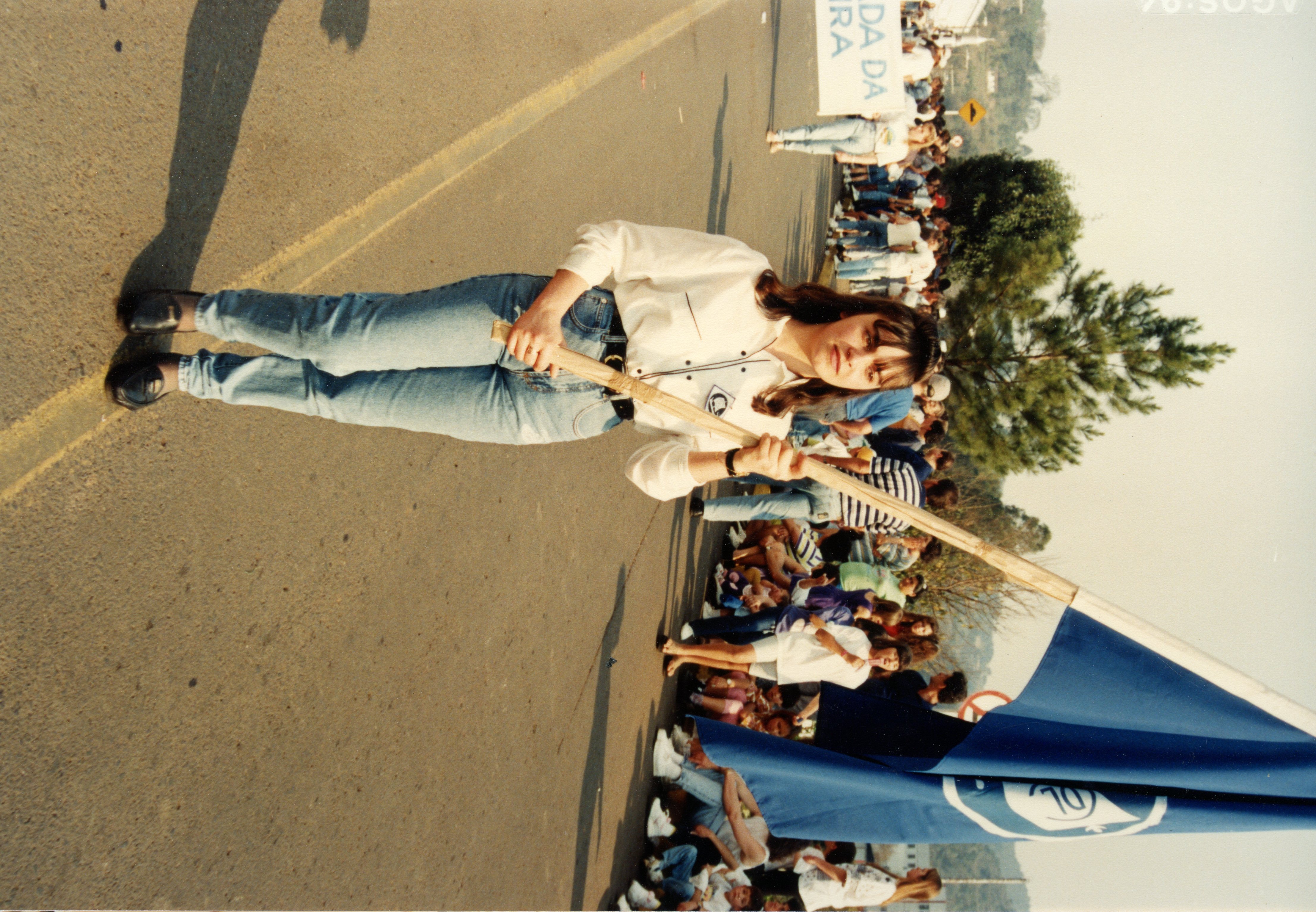 Uma jovem mulher segura uma bandeira azul durante um desfile ou evento público ao ar livre, vestindo camisa clara e calça jeans, típicas da moda dos anos 90. Ao fundo, observa-se uma multidão participando do evento em uma rua, com uma faixa parcialmente visível contendo a palavra 'IRA', possivelmente parte de 'VIDEIRA'.