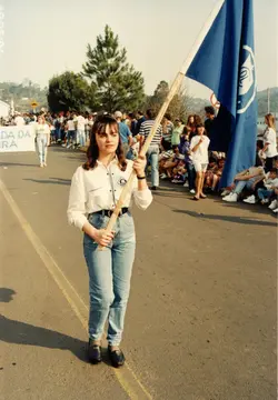 Uma jovem mulher segura uma bandeira azul durante um desfile ou evento público ao ar livre, vestindo camisa clara e calça jeans, típicas da moda dos anos 90. Ao fundo, observa-se uma multidão participando do evento em uma rua, com uma faixa parcialmente visível contendo a palavra 'IRA', possivelmente parte de 'VIDEIRA'.