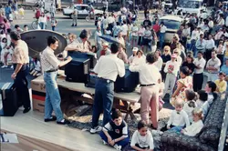 Uma cena ao ar livre em uma praça ou evento público, provavelmente na década de 1990, onde pessoas se reúnem em torno de televisores Toshiba expostos em mesas. O ambiente sugere uma demonstração tecnológica ou sorteio em uma cidade brasileira, com vestimentas típicas da época, como jeans de cintura alta e camisas polo.