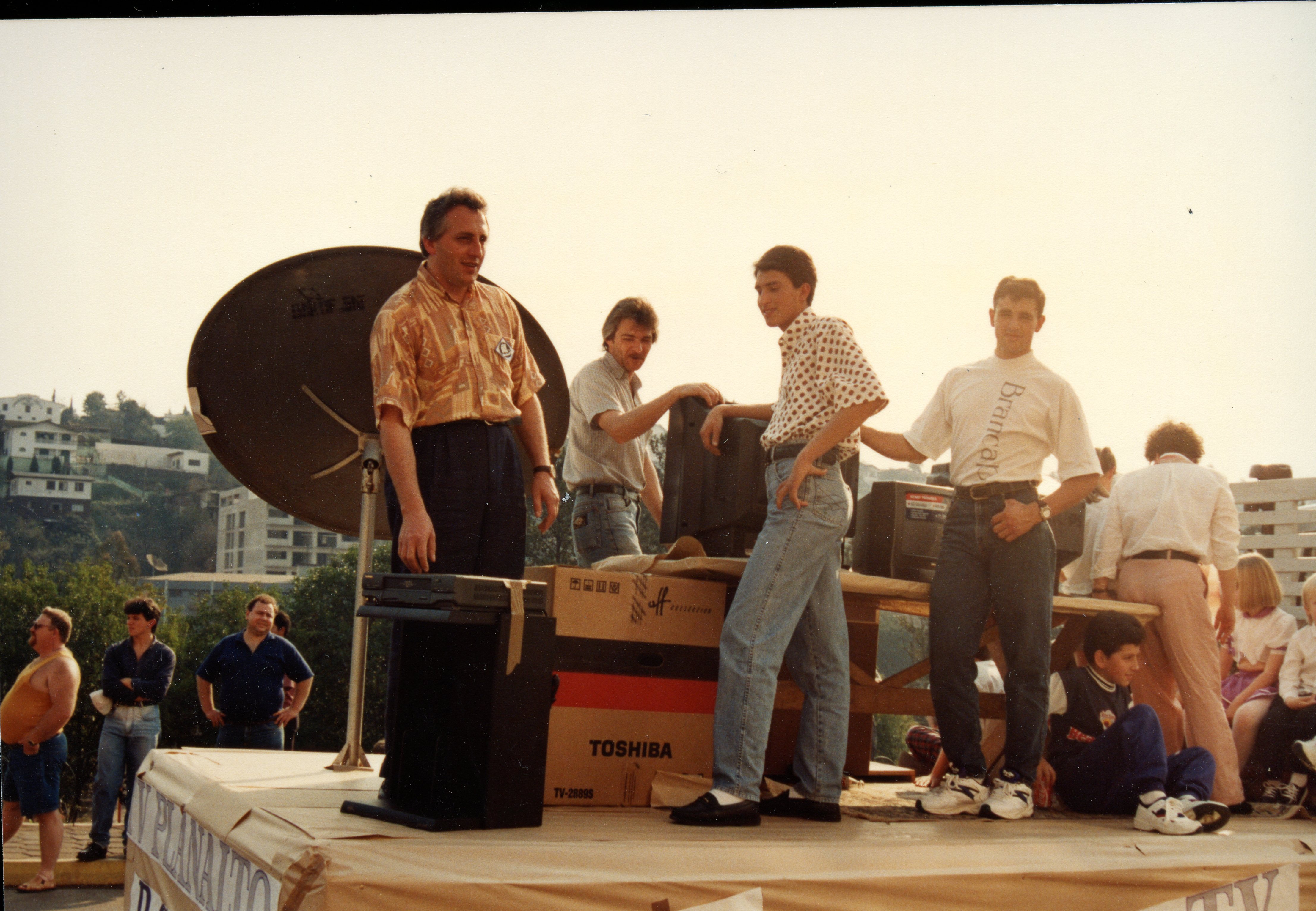 Uma fotografia colorida que retrata um grupo de homens em um palco ou plataforma ao ar livre, possivelmente durante um evento promocional ou feira de eletrônicos. Destacam-se equipamentos como uma antena parabólica e caixas de televisão da marca Toshiba, com vestimentas típicas da década de 1990, como camisas estampadas e jeans de cintura alta.