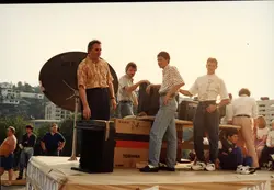 Uma fotografia colorida que retrata um grupo de homens em um palco ou plataforma ao ar livre, possivelmente durante um evento promocional ou feira de eletrônicos. Destacam-se equipamentos como uma antena parabólica e caixas de televisão da marca Toshiba, com vestimentas típicas da década de 1990, como camisas estampadas e jeans de cintura alta.