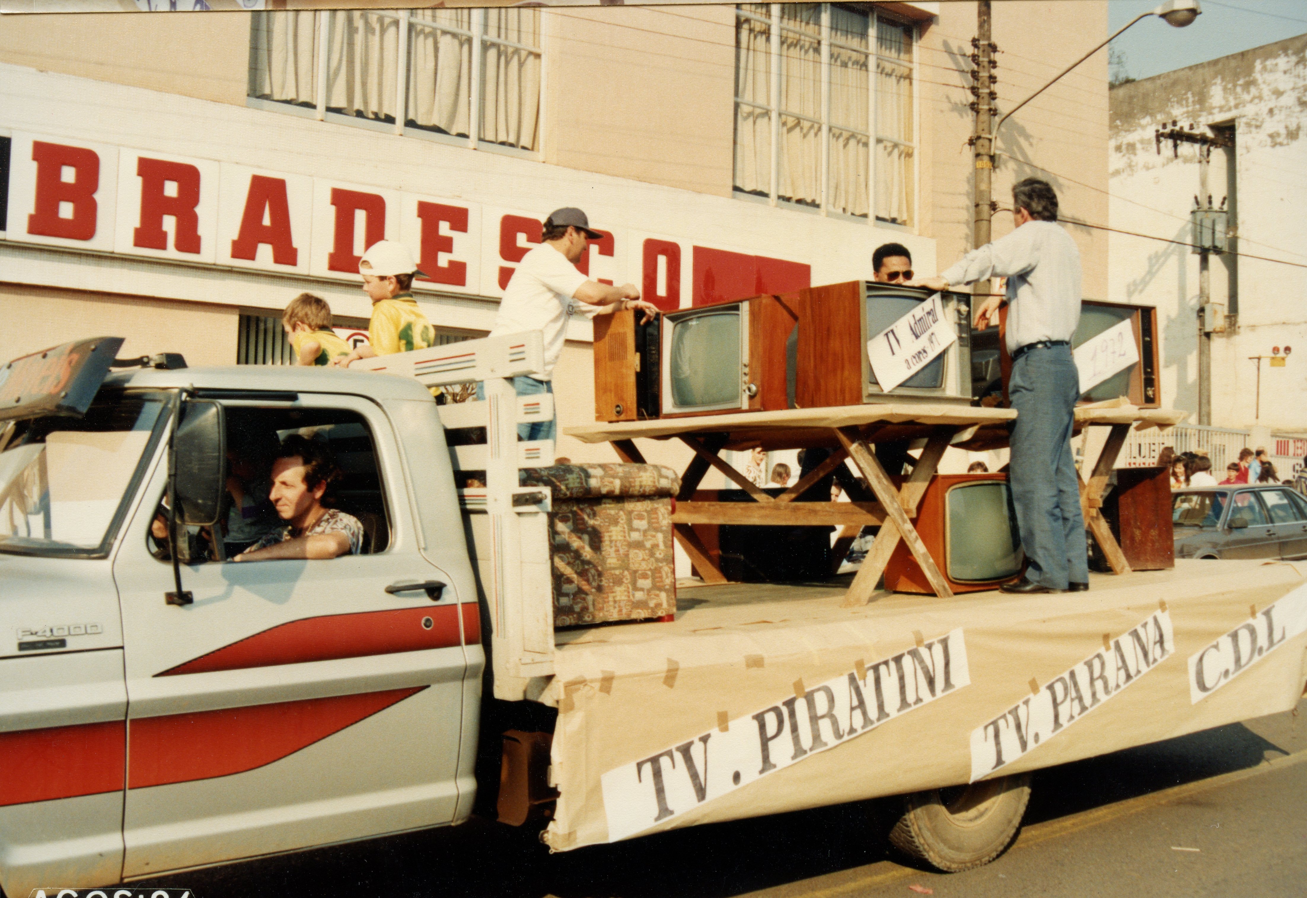 Uma fotografia de um desfile ou evento público em frente a uma agência do banco Bradesco, onde um caminhão transporta televisores antigos de tubo. O veículo exibe faixas com nomes de emissoras como TV Piratini e TV Paraná, sugerindo uma celebração histórica ou comemorativa ocorrida provavelmente na década de 1990.