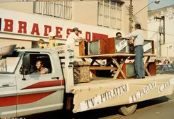 Uma fotografia de um desfile ou evento público em frente a uma agência do banco Bradesco, onde um caminhão transporta televisores antigos de tubo. O veículo exibe faixas com nomes de emissoras como TV Piratini e TV Paraná, sugerindo uma celebração histórica ou comemorativa ocorrida provavelmente na década de 1990.