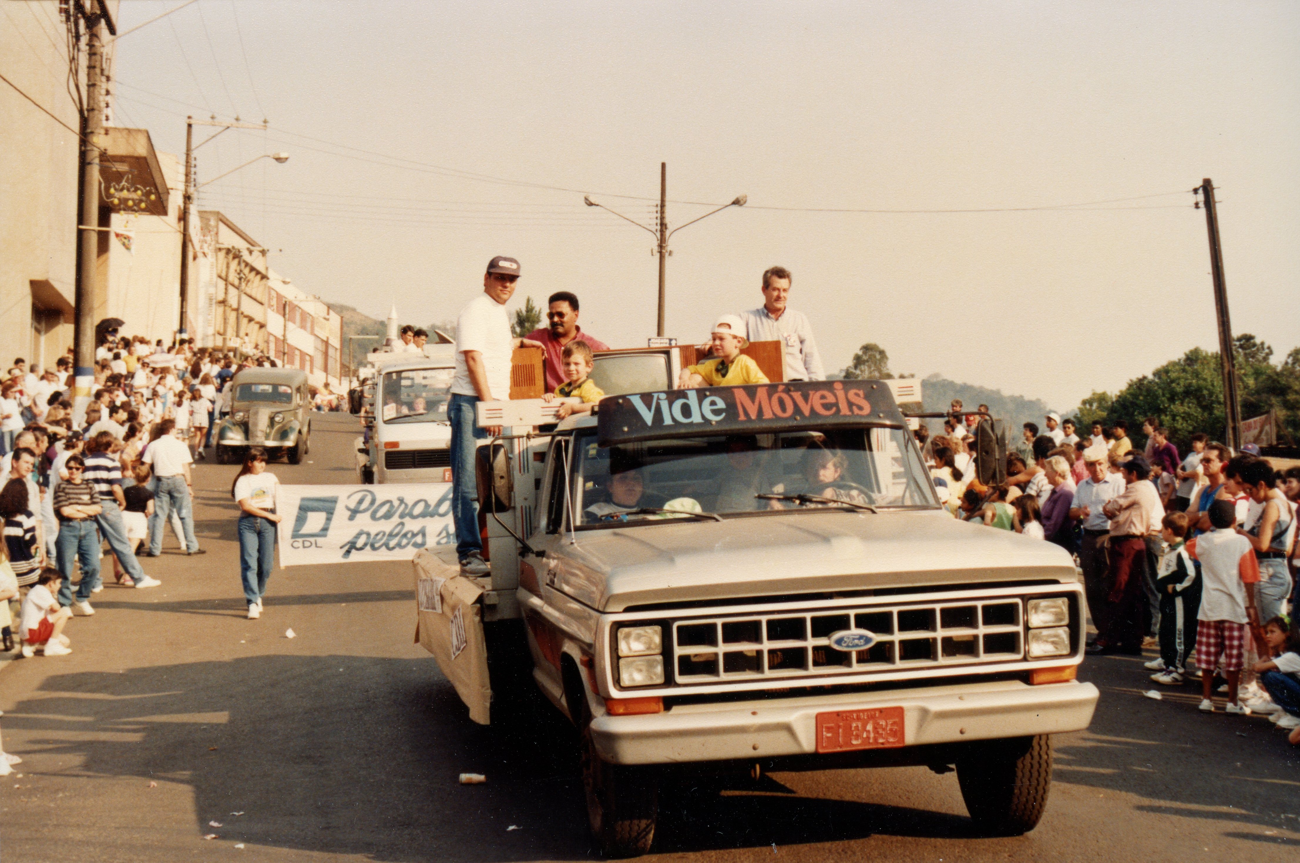 Uma fotografia de um desfile de rua em Videira, apresentando um caminhão Ford decorado da empresa 'Vide Móveis' com pessoas na carroceria. O evento parece ser uma celebração pública ou comemoração cívica, com uma multidão acompanhando o veículo em uma via urbana.