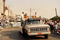 Uma fotografia de um desfile de rua em Videira, apresentando um caminhão Ford decorado da empresa 'Vide Móveis' com pessoas na carroceria. O evento parece ser uma celebração pública ou comemoração cívica, com uma multidão acompanhando o veículo em uma via urbana.