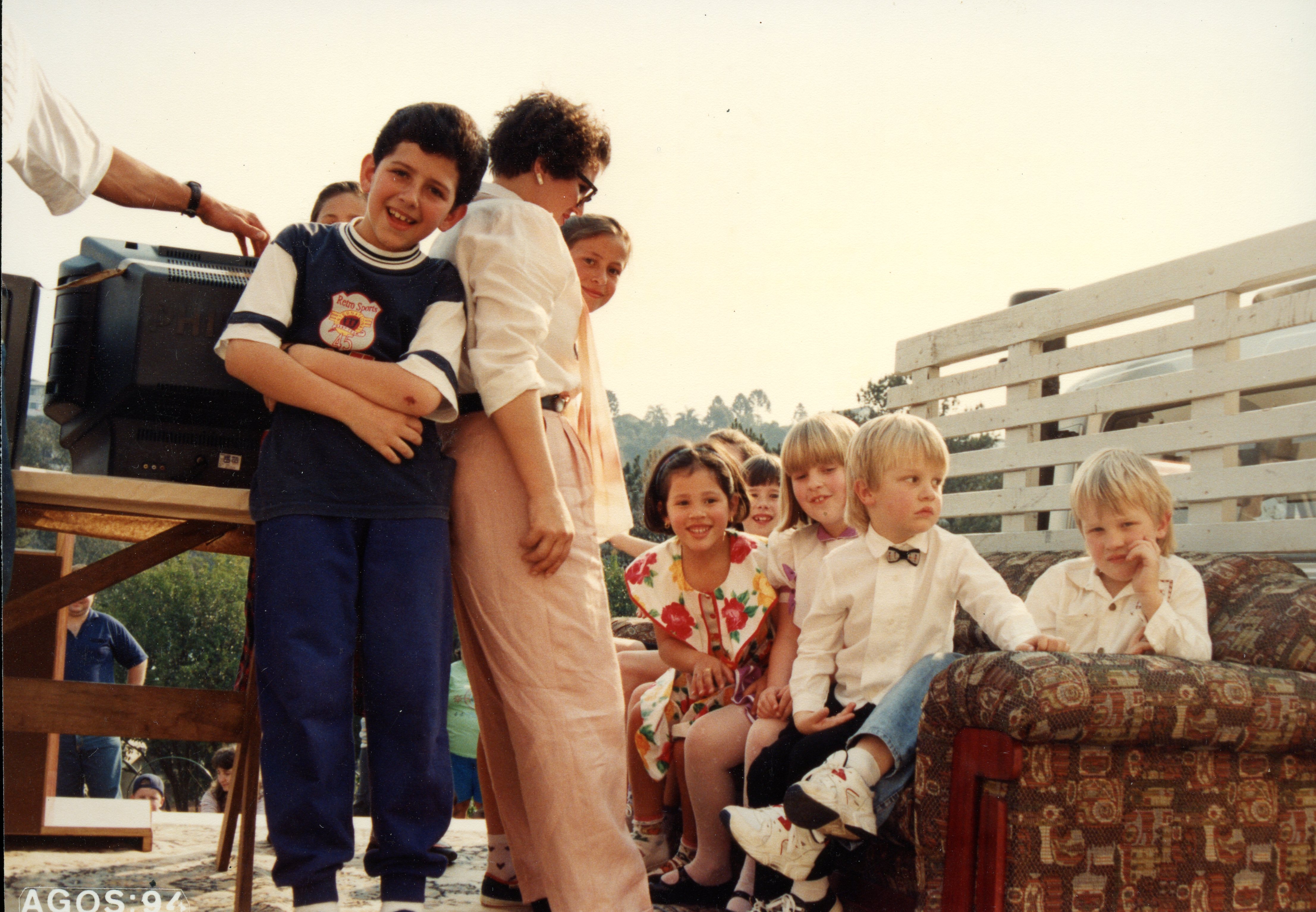 Uma fotografia colorida que captura um grupo de crianças e uma mulher em um ambiente externo, possivelmente durante uma celebração ou evento comunitário. A imagem apresenta vestimentas típicas da década de 1990, com crianças sentadas em um sofá colocado ao ar livre e uma televisão sobre uma mesa de madeira, sugerindo uma atividade recreativa ou educativa.