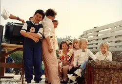 Uma fotografia colorida que captura um grupo de crianças e uma mulher em um ambiente externo, possivelmente durante uma celebração ou evento comunitário. A imagem apresenta vestimentas típicas da década de 1990, com crianças sentadas em um sofá colocado ao ar livre e uma televisão sobre uma mesa de madeira, sugerindo uma atividade recreativa ou educativa.